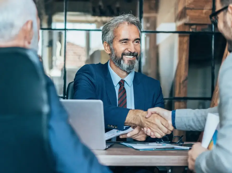 A smiling financial advisor shaking hands with a small business client across a desk with financial documents and a laptop, representing an outsourced CFO partnership and strategic business advisory.