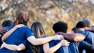 A diverse group of volunteers in matching blue shirts stand together with arms around each other, representing community partnership and local business engagement.