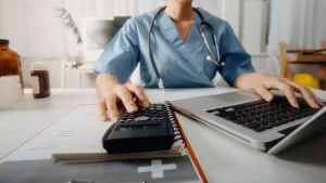 A healthcare professional in scrubs uses a calculator and laptop at a desk with medical documents, representing tax planning strategies for medical practices and healthcare providers.